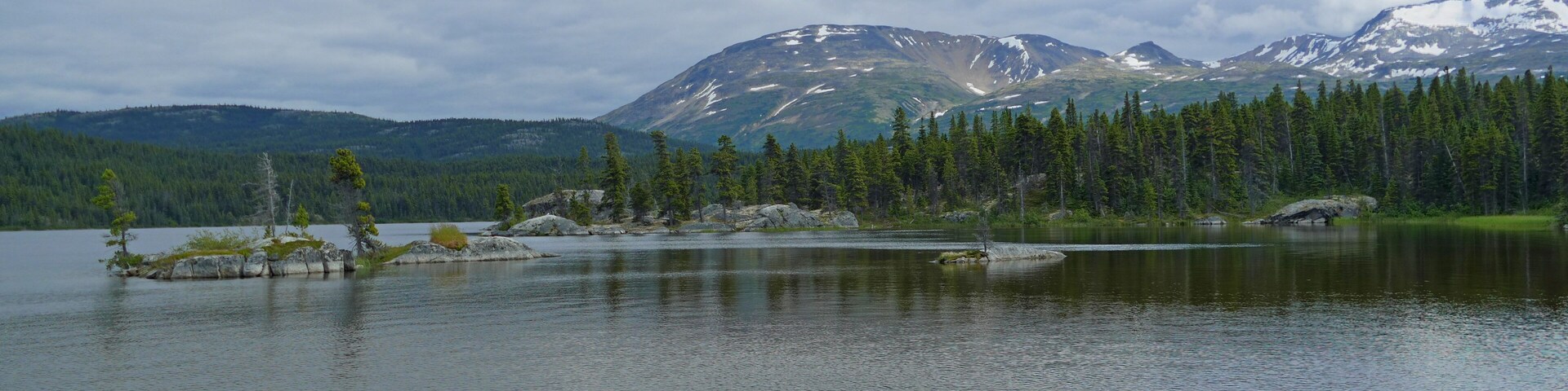 A calm lake surrounded by dense forests and snow-capped mountains under a cloudy sky.