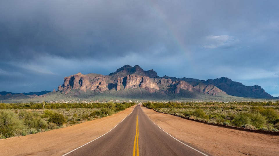 A road leading to a mountain range with a rainbow in the sky.
