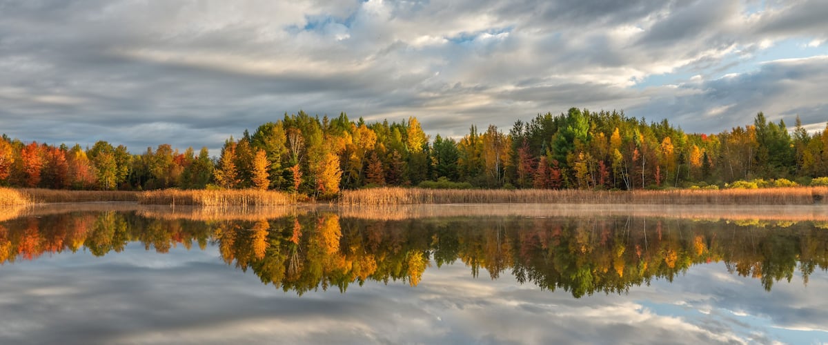 A calm lake reflecting autumn trees and a cloudy sky.