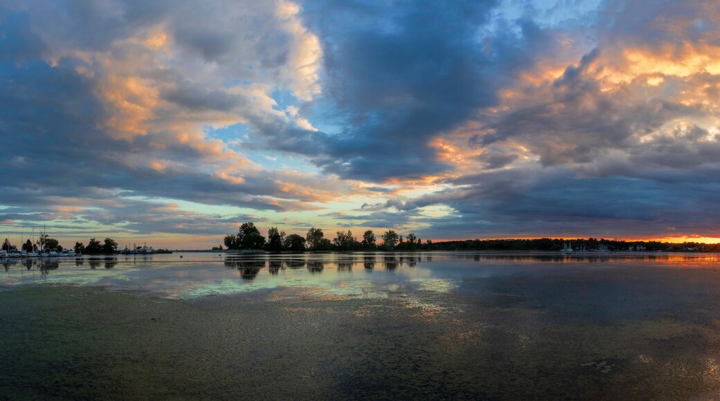 A sunset over a calm lake with boats and trees reflecting on the water.
