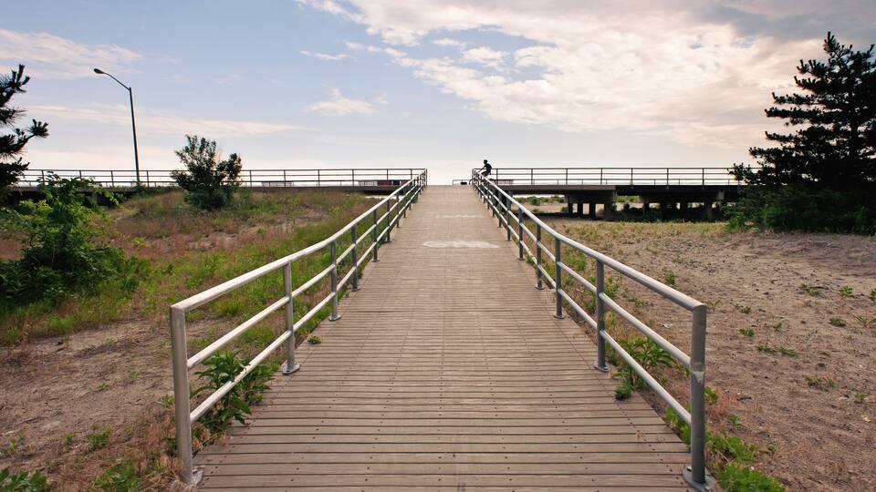 A wooden boardwalk with metal railings leading to a bridge over a body of water.