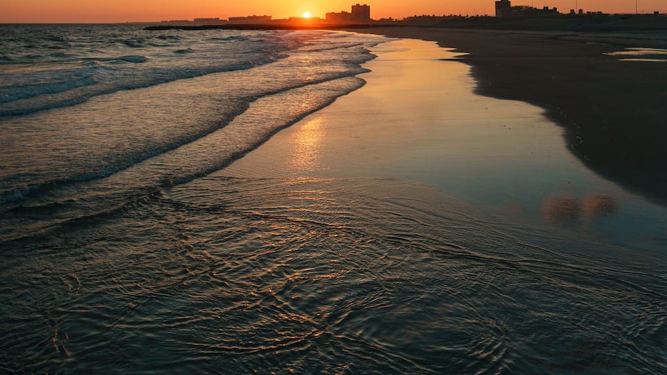 A beach at sunset with waves, a clear sky, and distant buildings.