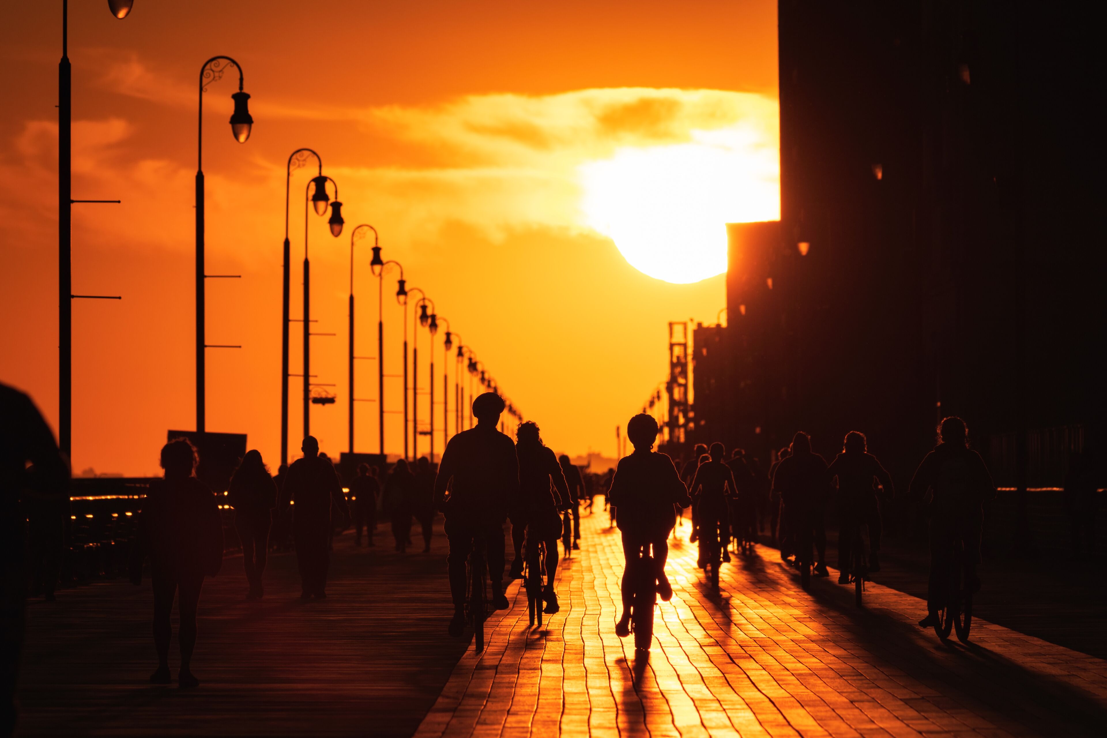 Large sun setting looking straight down a long boardwalk, with silhouettes of walkers and cyclists. Long Beach New York.