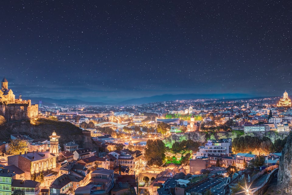 Tbilisi, Georgia. Panorama Of Narikala Fortress, Sameba Holy Trinity Cathedral, Old Historic District Abanotubani In Night Illumination Under Amazing Blue Bright Night Starry Sky.