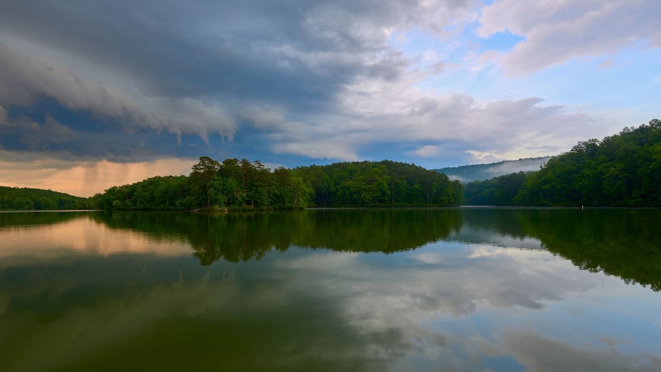 A calm lake with a wooden dock, surrounded by dense green forest and hills under a cloudy sky.