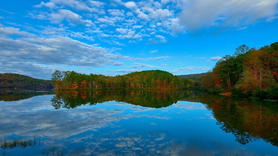 A calm lake reflecting trees with autumn foliage, surrounded by hills under a partly cloudy sky.