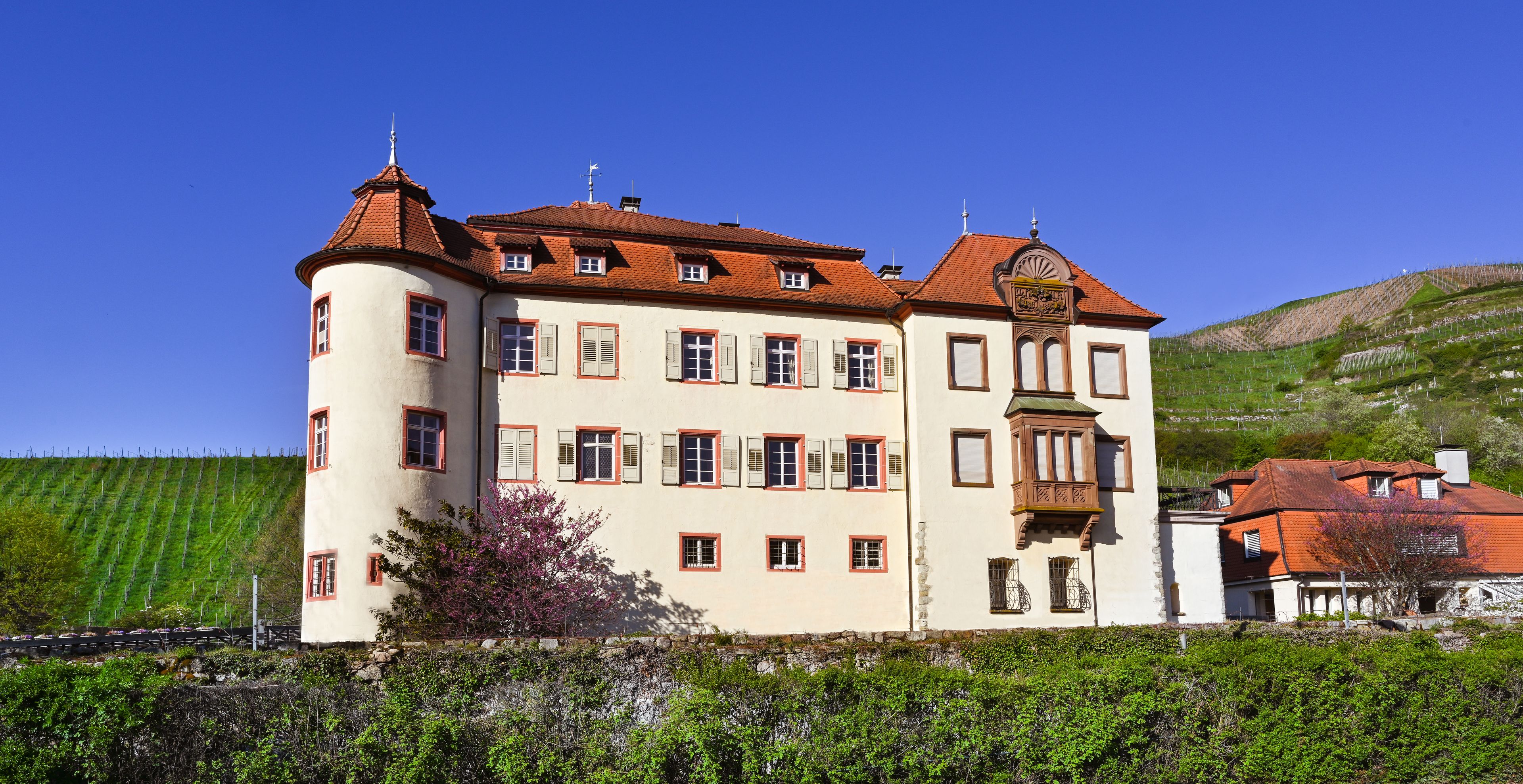 A large, multi-story building with a red-tiled roof, surrounded by lush greenery and a hillside.