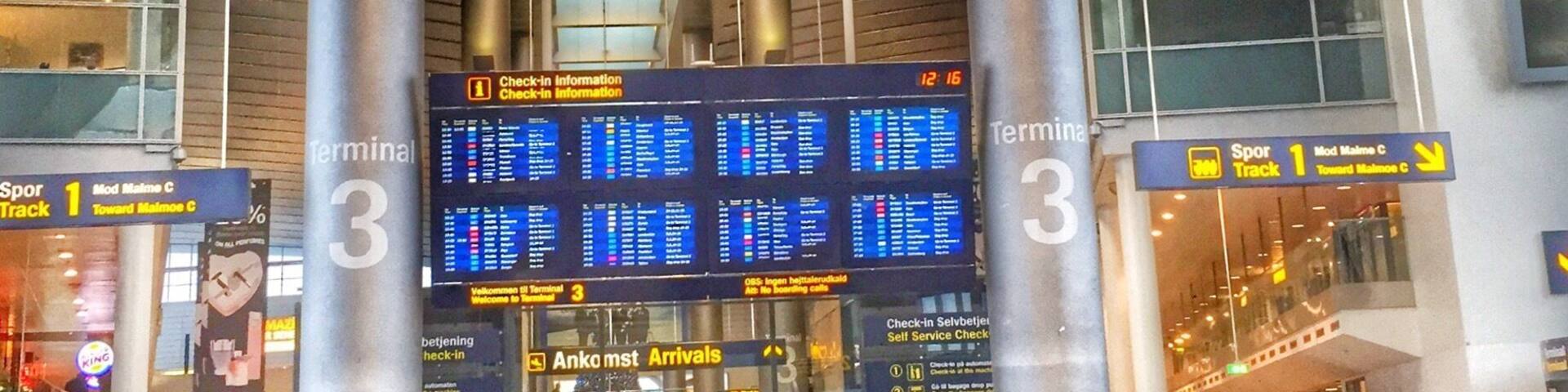 A busy airport terminal with a large digital flight information board, a clock, and people standing in line.