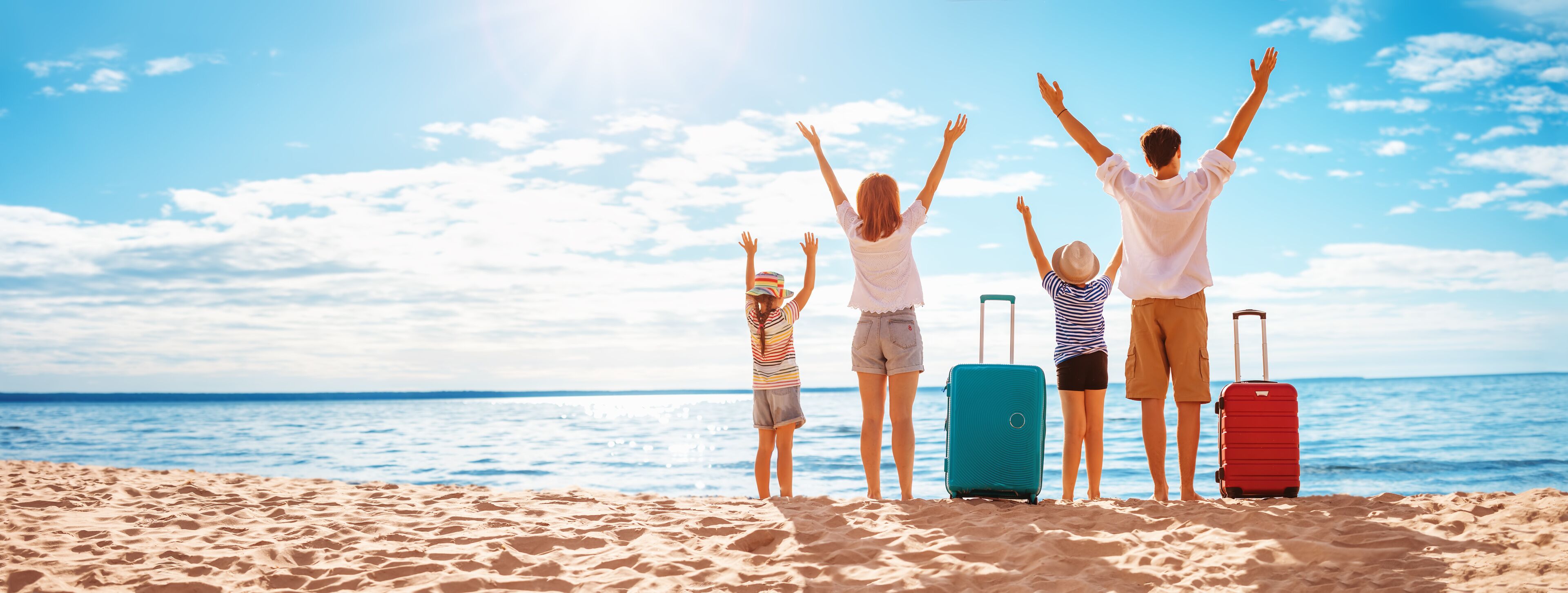 A beach with people standing and facing the ocean, with their arms raised. There are two suitcases on the sand.