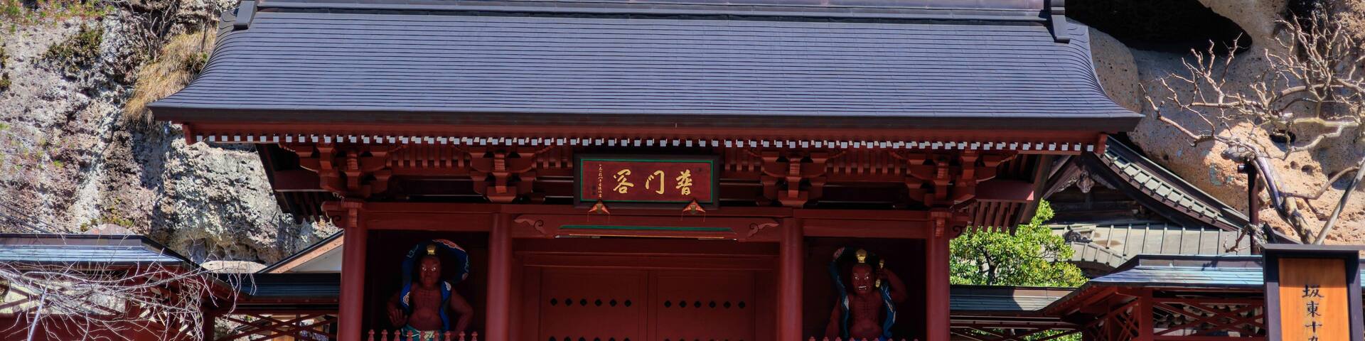 A traditional Japanese temple with a red entrance gate, stone steps leading up to it, and a rocky mountain backdrop.