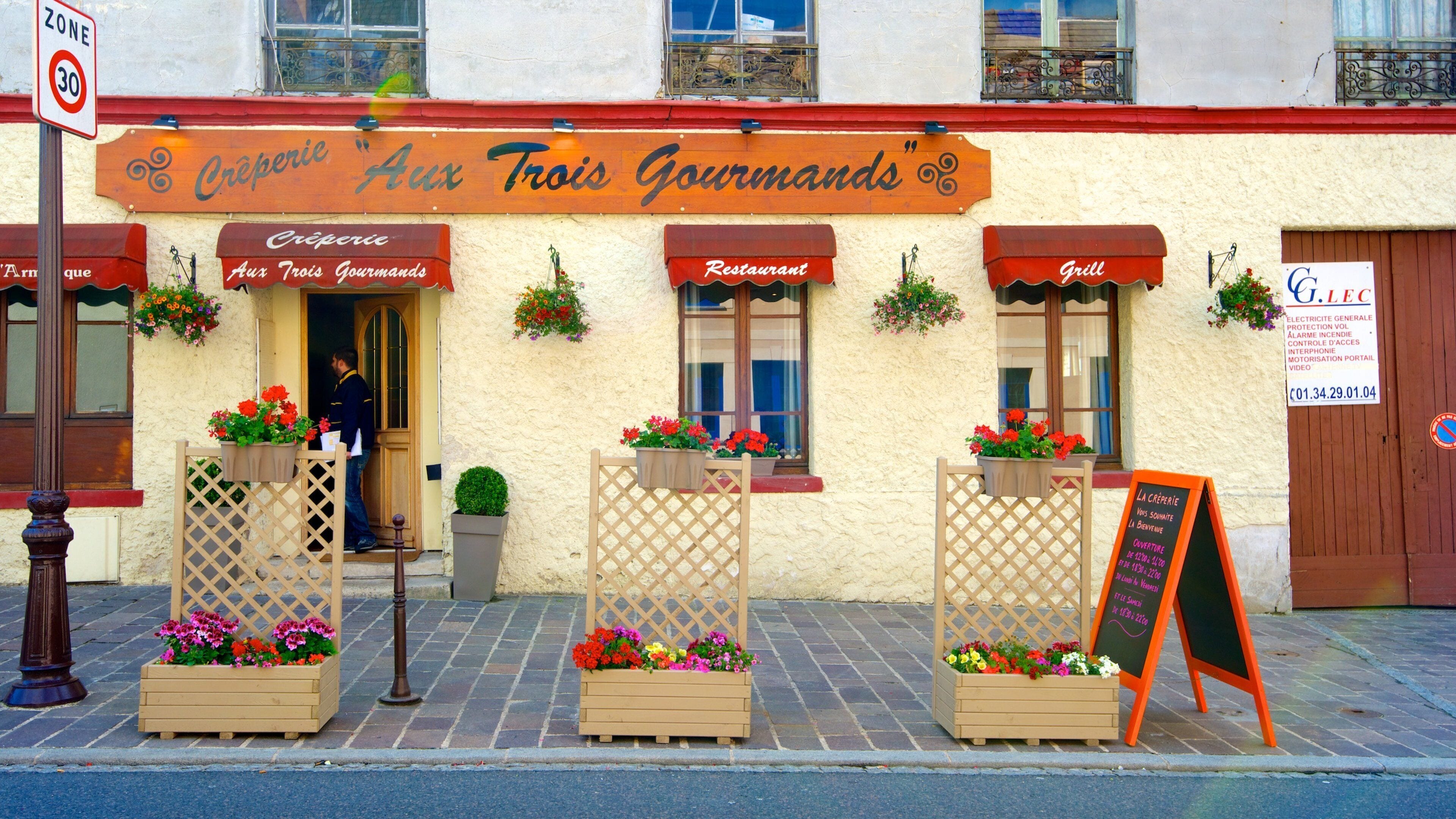 Un restaurante llamado "Crêperie Aux Trois Gourmands" con terraza y macetas con flores.