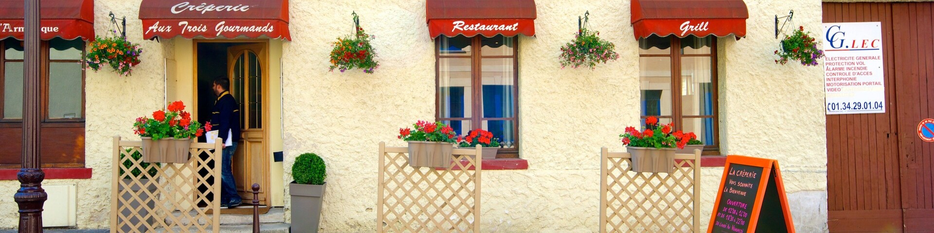 Un restaurante llamado "Crêperie Aux Trois Gourmands" con terraza y macetas con flores.