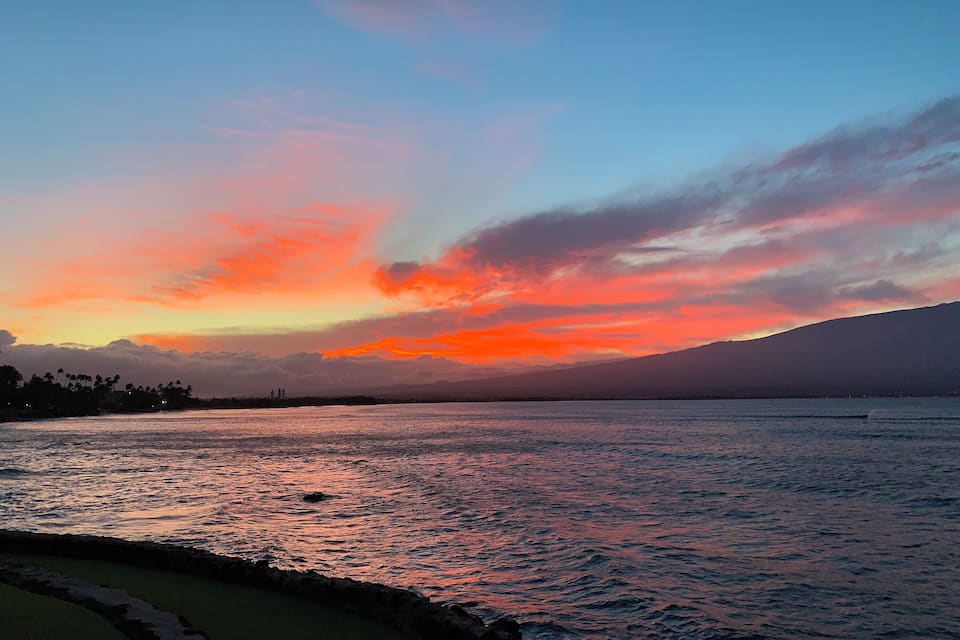 A sunset over a calm sea with a rocky shoreline and distant mountains.