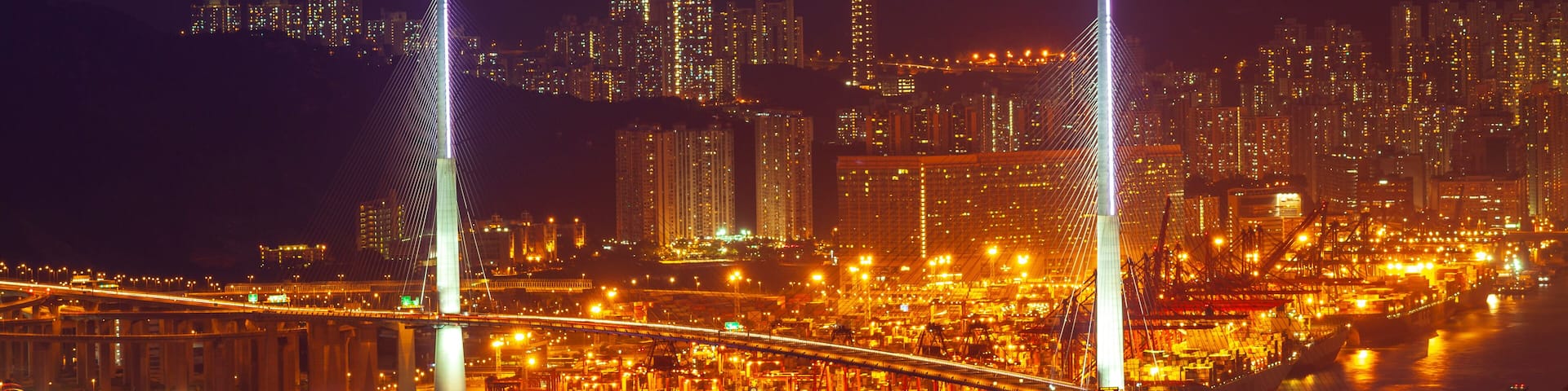 A nighttime cityscape with illuminated skyscrapers, a prominent cable-stayed bridge, and a busy harbor with ships.