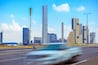 Stadsgezicht met moderne wolkenkrabbers, een hoog monument en een rijdend voertuig op de weg.