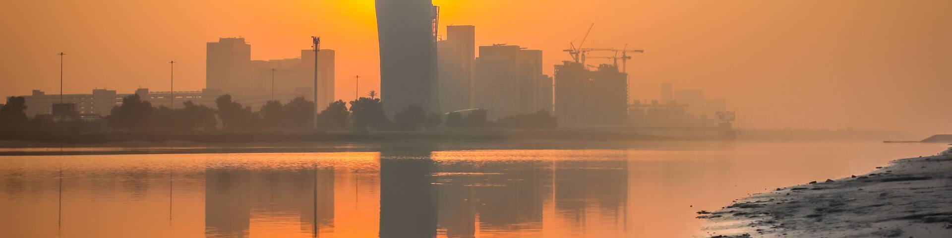 A sunrise over a city skyline reflected in calm waters, with buildings and construction cranes visible.