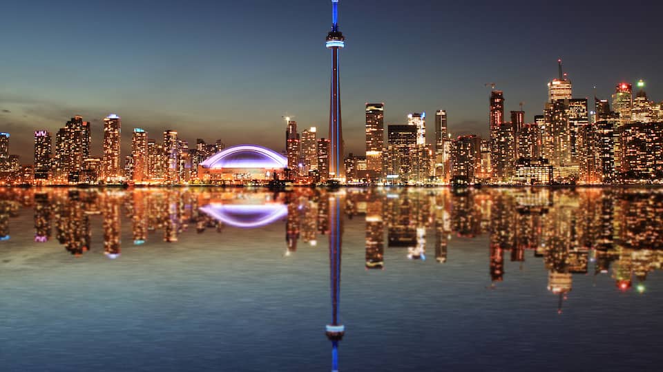 A city skyline at night with a prominent tower and a stadium reflecting on the water.