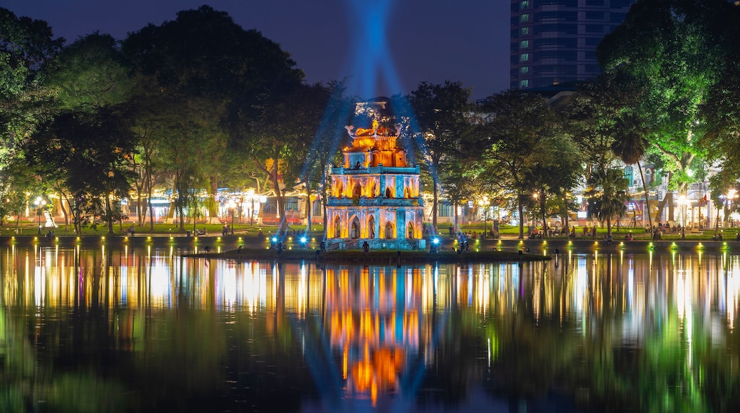 A brightly lit pagoda with reflections in a calm body of water, surrounded by trees and a cityscape in the background.