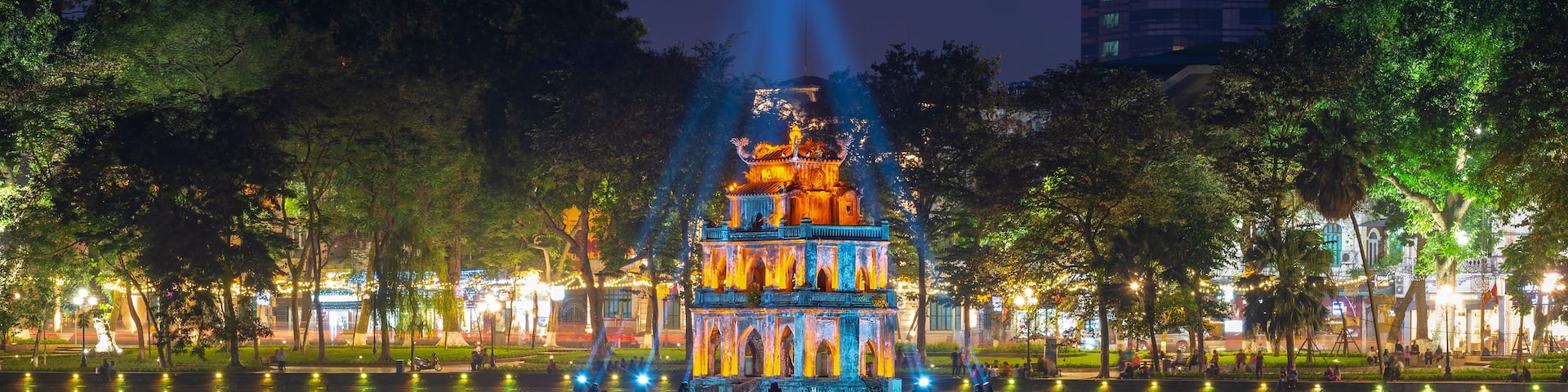A brightly lit pagoda with reflections in a calm body of water, surrounded by trees and a cityscape in the background.