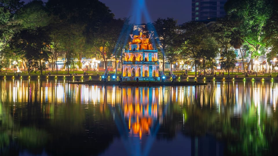 Um pagode bem iluminado com reflexos em águas calmas, cercado por árvores e um horizonte urbano ao fundo.
