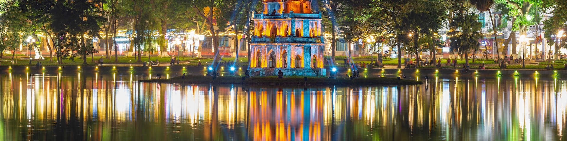A brightly lit pagoda with reflections in a calm body of water, surrounded by trees and a cityscape in the background.