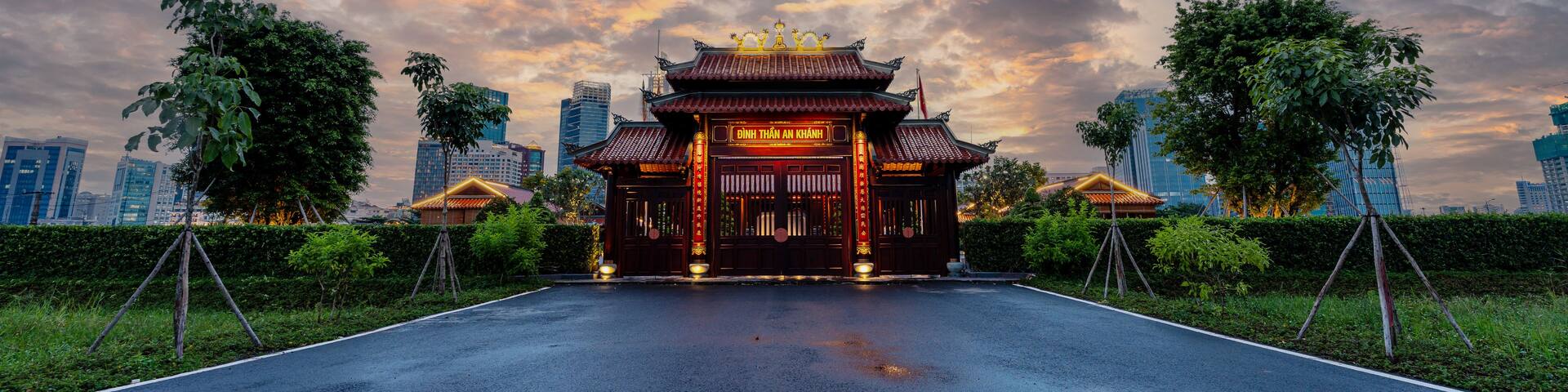 A traditional building with a golden roof and red walls, surrounded by greenery and modern buildings in the background.