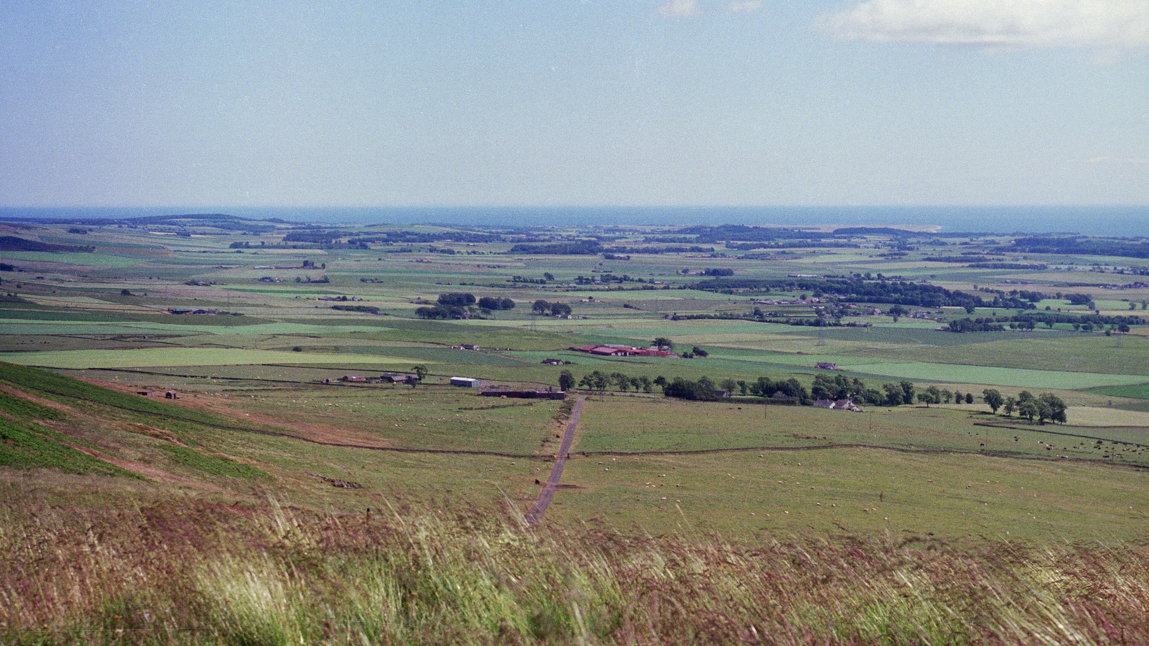 A rural landscape with rolling hills, scattered fields, and a few small buildings.