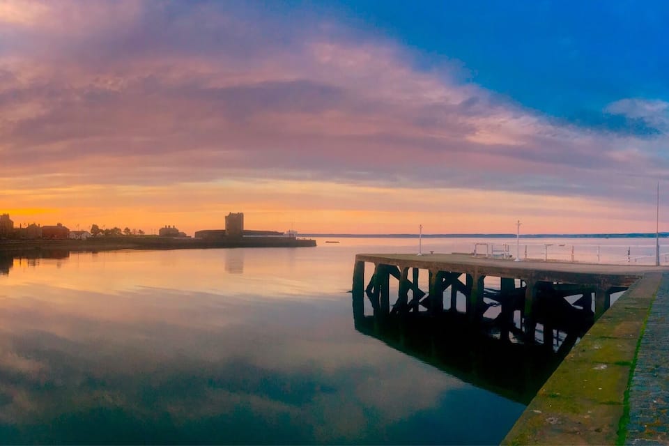 Een zonsondergang boven een rustige rivier met een steiger en gebouwen op de achtergrond.