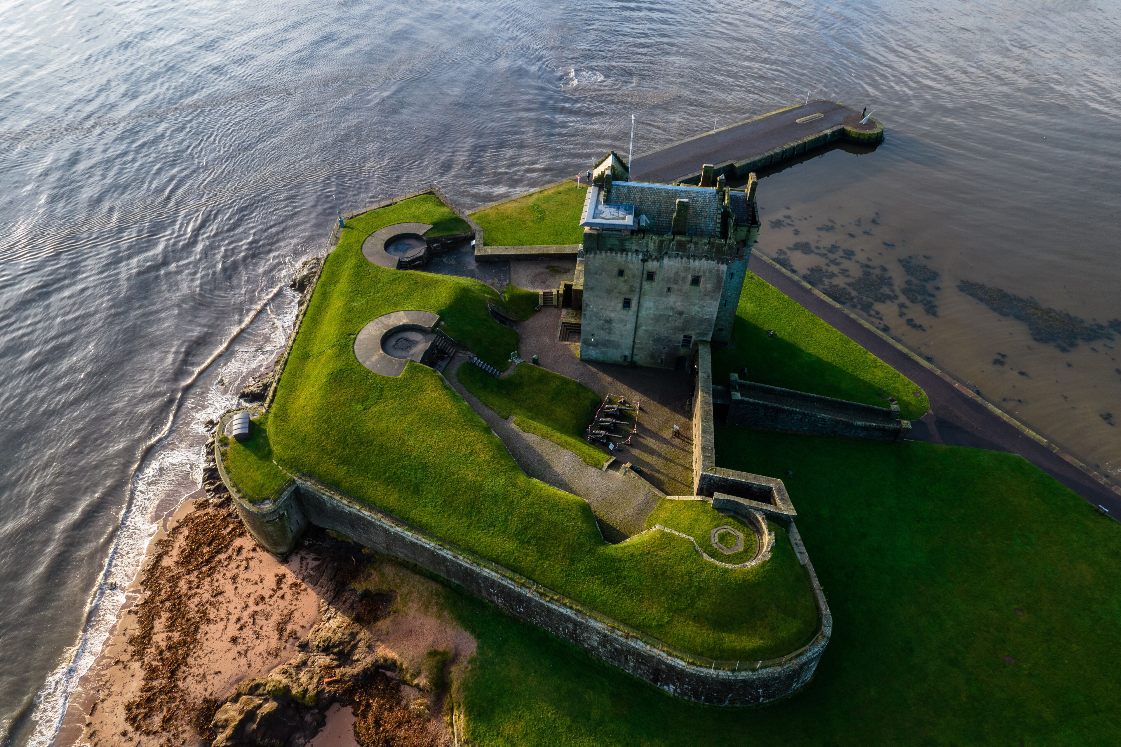 An aerial view of a coastal fort with a grassy embankment, a circular water feature, and a large tower.
