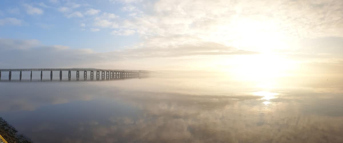 A bridge extending over calm water with a sunrise or sunset in the background.