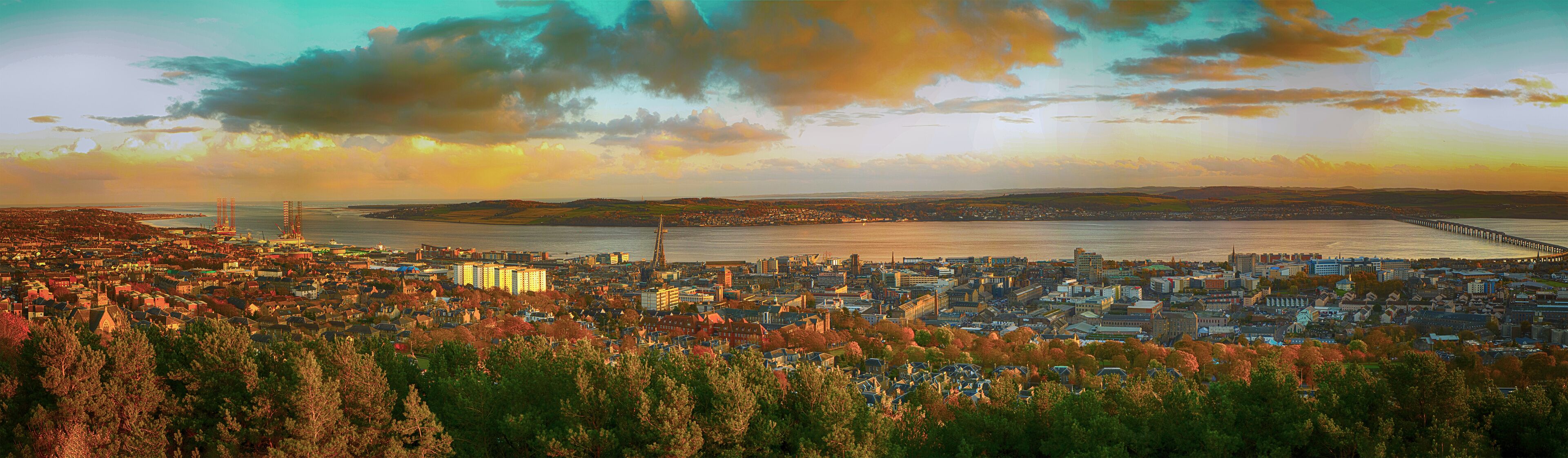 A coastal cityscape with a prominent pier extending into the water, surrounded by dense urban buildings and a backdrop of hills.