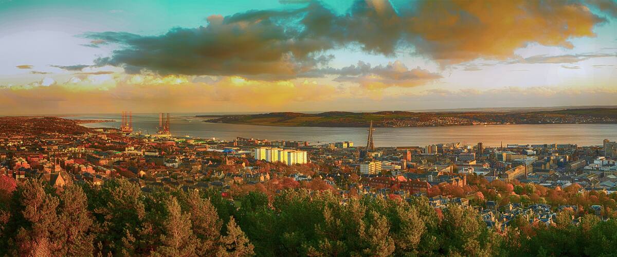 A coastal cityscape with a prominent pier extending into the water, surrounded by dense urban buildings and a backdrop of hills.
