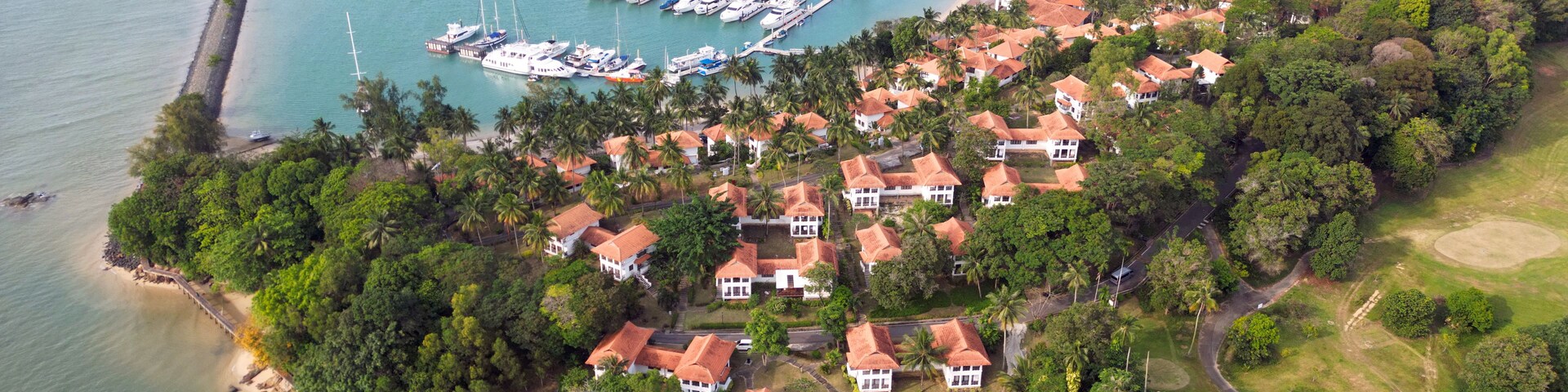 An aerial view of a coastal town with a marina, houses with red roofs, and a bridge connecting the land to the water.