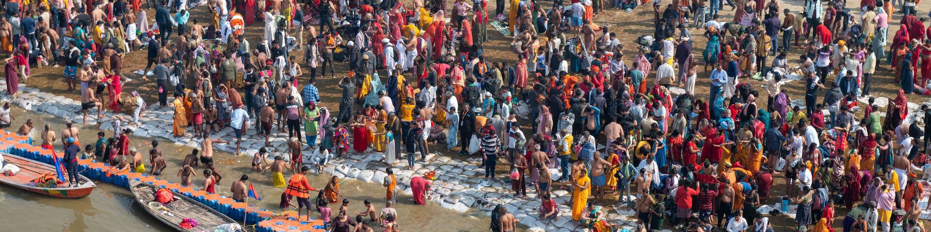 A crowded riverside with people bathing and boats lined up along the shore.