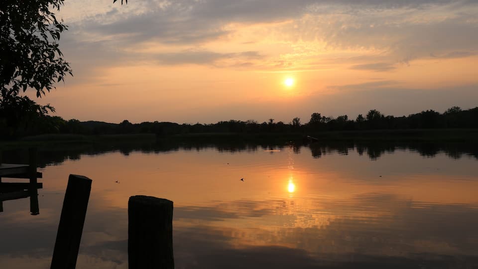 A sunset over a calm lake with reflections of the sky, surrounded by trees and a dock.