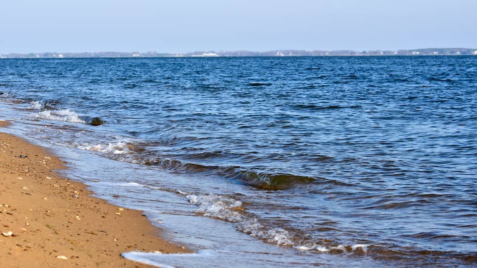 A sandy beach with gentle waves and a distant shoreline with buildings.