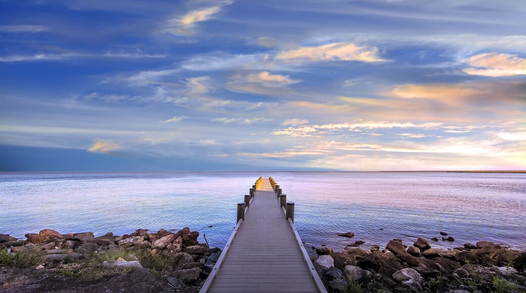 A wooden pier leading into a calm sea with a rocky shoreline and a cloudy sky.