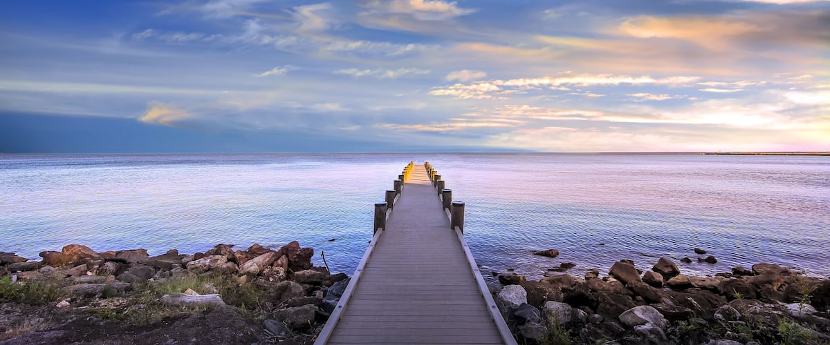 A wooden pier leading into a calm sea with a rocky shoreline and a cloudy sky.