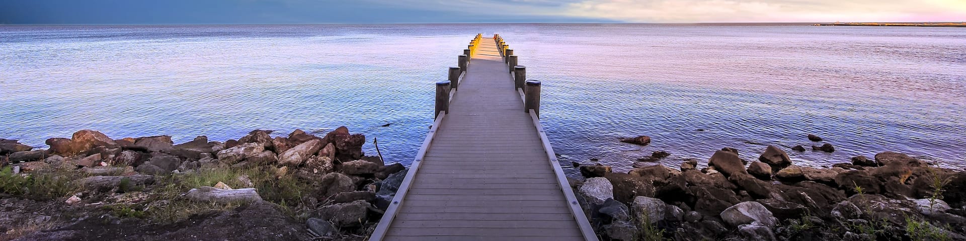 A wooden pier leading into a calm sea with a rocky shoreline and a cloudy sky.