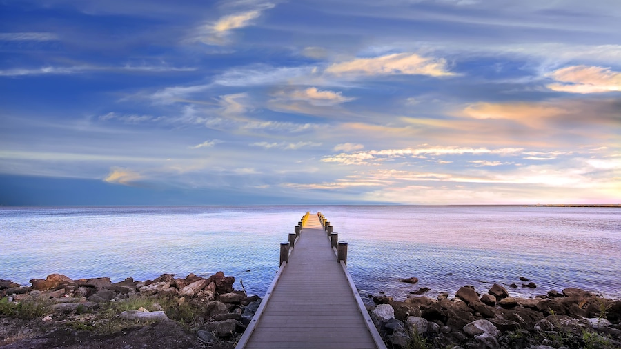 Un muelle de madera que se adentra en un mar tranquilo, con una costa rocosa y un cielo nublado.