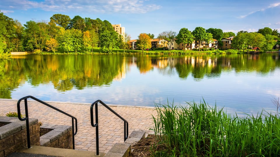A calm lake with a reflection of trees and buildings, surrounded by greenery and a paved walkway.