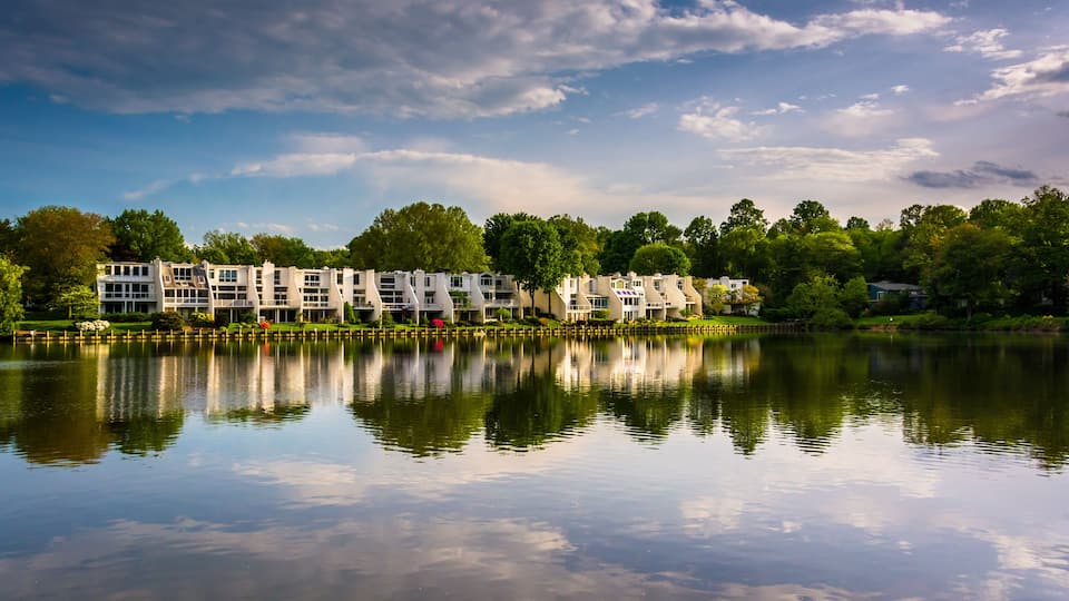 A calm lake reflecting buildings and trees under a partly cloudy sky.
