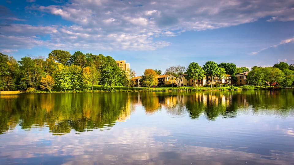 A calm lake with reflections of trees and buildings, surrounded by greenery and a partly cloudy sky.