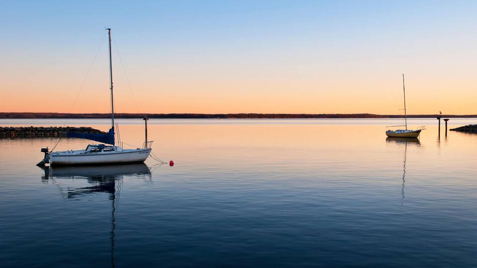 A calm lake with two sailboats anchored near a rocky shore at sunset.