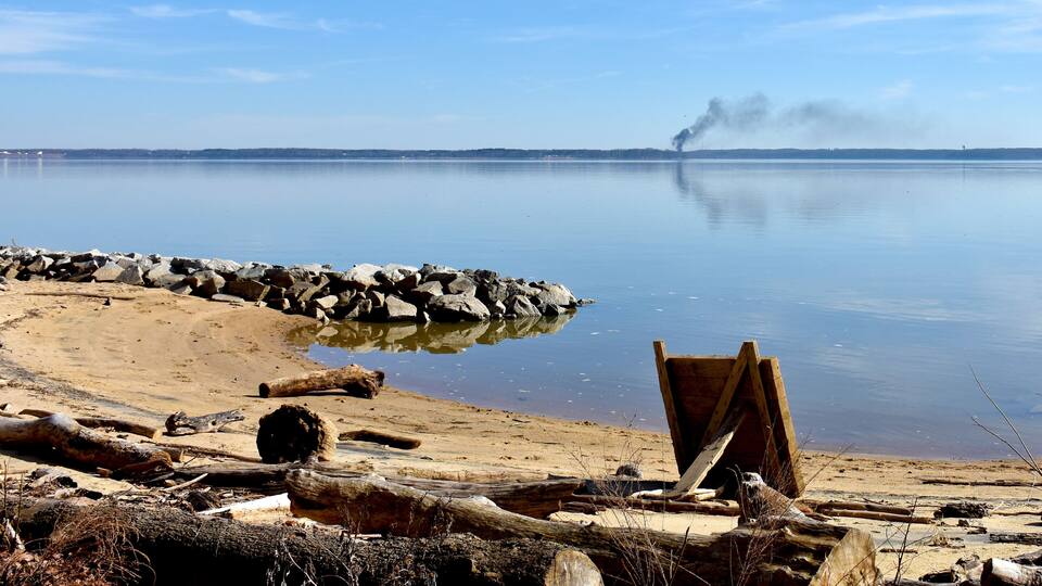 A calm lake with a rocky shoreline, a wooden structure, and a distant smokestack.