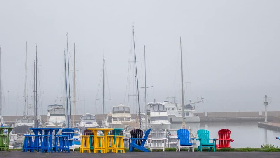 A marina with multiple sailboats docked, colorful Adirondack chairs, and a foggy atmosphere.
