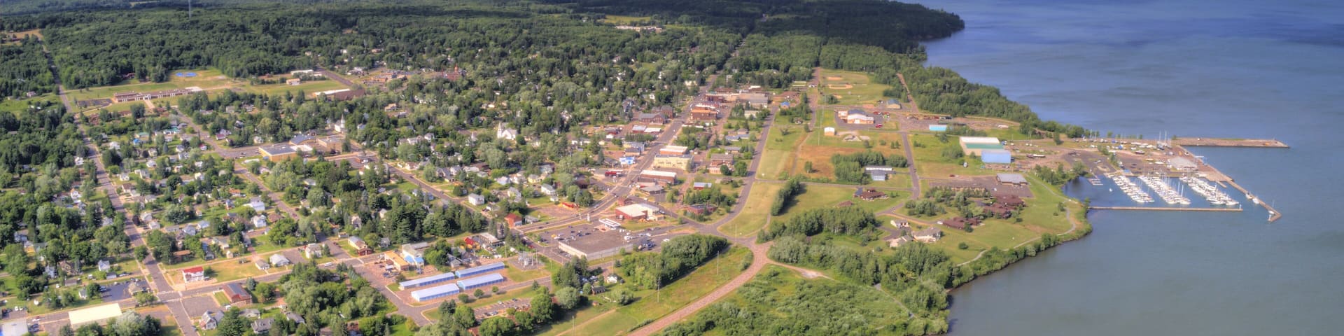 An aerial view of a coastal town with a marina, dense forest, and a clear blue sky.
