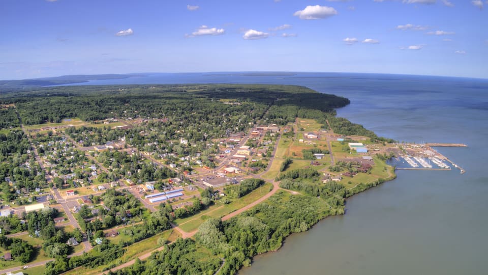 An aerial view of a coastal town with a marina, dense forest, and a clear blue sky.