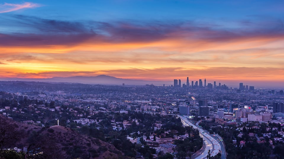 A cityscape at sunset with a prominent skyline, a winding road, and a hill in the foreground.
