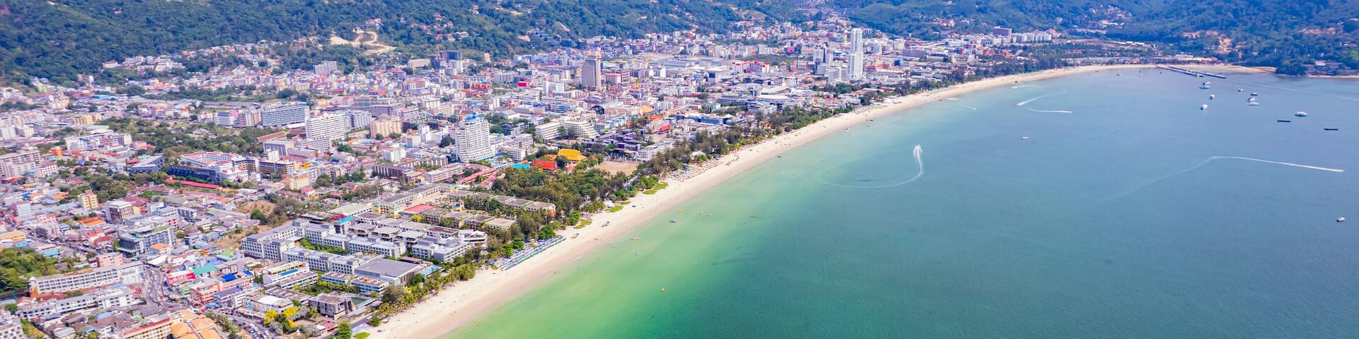 A coastal city with a long sandy beach, clear turquoise waters, and a backdrop of green hills.